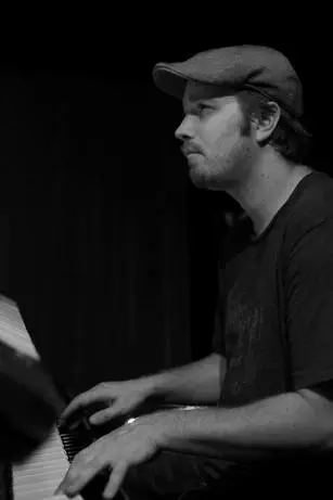 A man in black shirt playing piano with hat on.