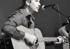 A man playing an acoustic guitar while holding a microphone.